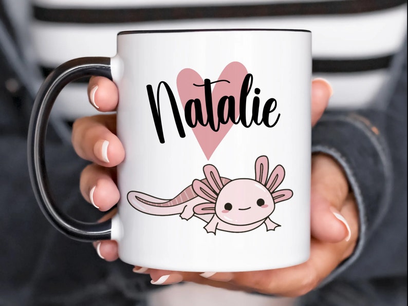 K&ouml;nnte beinhalten: Wei&szlig;er Keramikbecher mit schwarzem Henkel und Rand. Der Becher zeigt den Namen "Natalie" in schwarzer Schrift &uuml;ber einem rosa Herz und eine Cartoon-Axolotl-Illustration in Rosa.