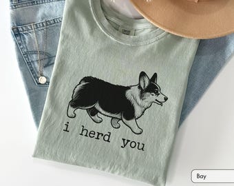 May include: A sage green t-shirt featuring a black and white illustration of a Corgi dog in profile. Below the dog, the text reads "i herd you" in a simple, lowercase black font. The shirt is displayed with light blue denim fabric.