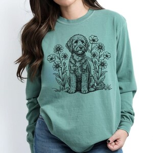 Pode incluir: Uma camisa de manga comprida verde claro com uma ilustra&ccedil;&atilde;o a preto e branco de um c&atilde;o sentado entre flores. A camisa &eacute; de cor lisa e tem decote redondo.