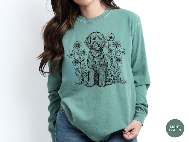 Pode incluir: Uma camisa de manga comprida verde claro com uma ilustra&ccedil;&atilde;o a preto e branco de um c&atilde;o sentado entre flores. A camisa &eacute; de cor lisa e tem decote redondo.