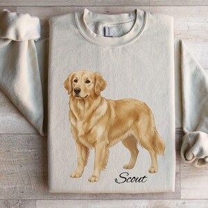 May include: Sand-colored sweatshirt featuring a detailed illustration of a Golden Retriever dog. The dog is standing, with the name "Scout" in script below. The sweatshirt has a crew neck and long sleeves.