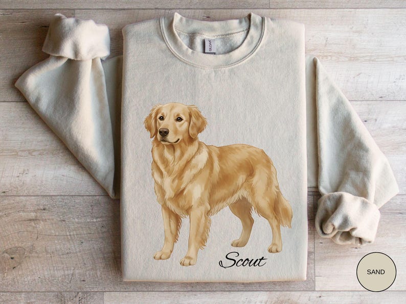 May include: Sand-colored sweatshirt featuring a detailed illustration of a Golden Retriever dog. The dog is standing, with the name "Scout" in script below. The sweatshirt has a crew neck and long sleeves.