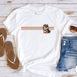 Puede incluir: Camiseta blanca con un gr&aacute;fico de perro pomerania marr&oacute;n y negro y un dise&ntilde;o de rayas horizontales en rosa, naranja y amarillo. La camiseta est&aacute; sobre una superficie de madera blanca con sandalias marrones y pantalones cortos de mezclilla azul.