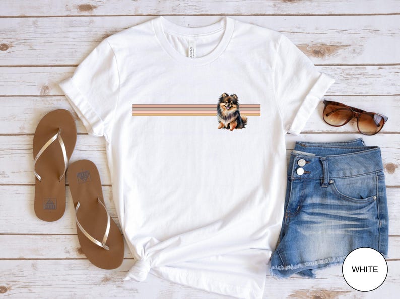 Puede incluir: Camiseta blanca con un gr&aacute;fico de perro pomerania marr&oacute;n y negro y un dise&ntilde;o de rayas horizontales en rosa, naranja y amarillo. La camiseta est&aacute; sobre una superficie de madera blanca con sandalias marrones y pantalones cortos de mezclilla azul.