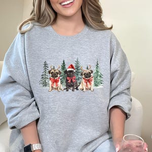 May include: A gray sweatshirt with a Christmas-themed design featuring three pugs wearing Santa hats and reindeer antlers. The pugs are standing in front of a snowy forest scene.