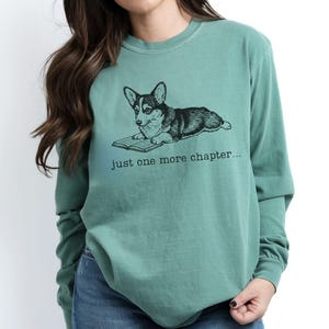 Op de afbeelding: Lichtgroen shirt met lange mouwen en een afbeelding van een corgi-hond die een boek leest. De tekst "just one more chapter..." staat onder de hond. Het shirt is gemaakt van een zacht materiaal.
