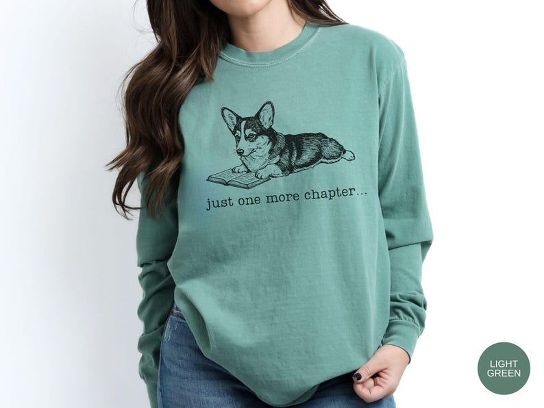 Op de afbeelding: Lichtgroen shirt met lange mouwen en een afbeelding van een corgi-hond die een boek leest. De tekst "just one more chapter..." staat onder de hond. Het shirt is gemaakt van een zacht materiaal.