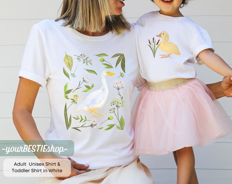 May include: White t-shirt with a watercolor illustration of a goose and floral details. The text "yourBESTIEShop" is printed on the bottom of the shirt.  Adult Unisex Shirt & Toddler Shirt in White.