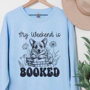 Puede incluir: Sudadera azul claro con un gr&aacute;fico de un perro corgi sentado en una pila de libros. El texto "My weekend is booked" est&aacute; impreso en la sudadera.