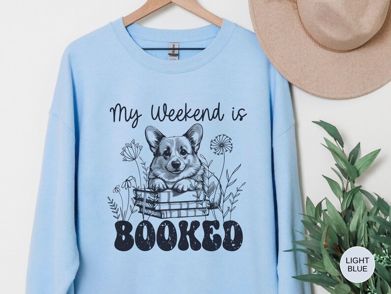 Puede incluir: Sudadera azul claro con un gr&aacute;fico de un perro corgi sentado en una pila de libros. El texto "My weekend is booked" est&aacute; impreso en la sudadera.