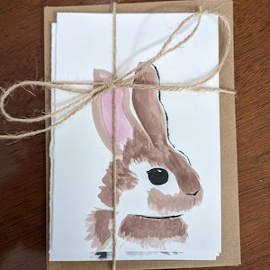 May include: A set of blank greeting cards featuring a watercolor illustration of a brown and white rabbit. The cards are tied with twine and packaged in a brown paper envelope. The rabbit has pink inner ears and a black eye.