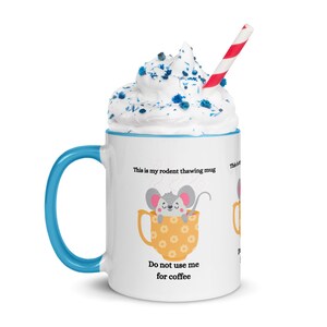 May include: A white ceramic mug with a blue handle and rim, topped with whipped cream and a red and white striped straw. The mug features a cartoon mouse in a yellow cup with the text "This is my rodent thawing mug" and "Do not use me for coffee."