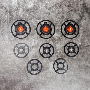 Op de afbeelding: Acht zwarte, ronde metalen doelen met de nummers 1, 2 en 3 in oranje vierkanten. De doelen zijn in een cirkel op een grijs betonnen oppervlak gerangschikt.