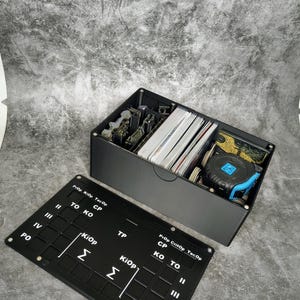 Könnte beinhalten: Eine schwarze Aufbewahrungsbox mit Deckel, die Karten, Miniaturen und ein Maßband enthält. Der Deckel ist schwarz mit weißem Text und Symbolen. Die Box steht auf einer grauen Oberfläche.