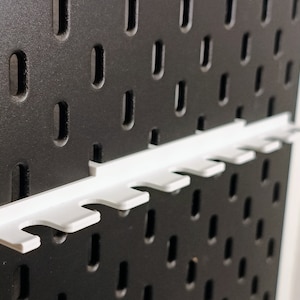 May include: White plastic pegboard organizer with six slots for holding tools or other items. The organizer is mounted on a black pegboard.