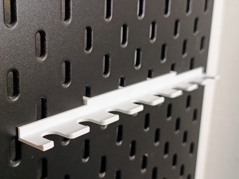 May include: White plastic pegboard organizer with six slots for holding tools or other items. The organizer is mounted on a black pegboard.