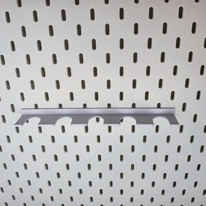 May include: A white pegboard with a gray metal pegboard hook with five slots. The hook is designed to hold tools or other items.