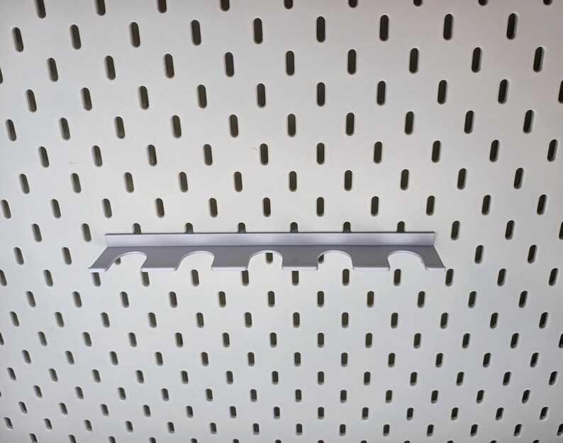 May include: A white pegboard with a gray metal pegboard hook with five slots. The hook is designed to hold tools or other items.