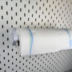 Könnte beinhalten: Eine weiße Küchenpapierrolle mit blau-grünen Streifen ist an einer weißen Lochwand befestigt. Die Küchenpapierrolle wird von einem weißen Kunststoffhalter gehalten.