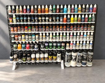 Könnte beinhalten: Ein schwarzes, mehrstufiges Präsentationsregal mit zahlreichen kleinen Kunststoffflaschen für Hobbyfarben in verschiedenen Farben. Das Regal verfügt über Reihen von kreisförmigen Aussparungen, um die Flaschen sicher zu halten.