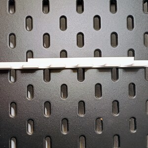 May include: White plastic pegboard hook strip with 10 hooks. The strip is mounted on a black pegboard.