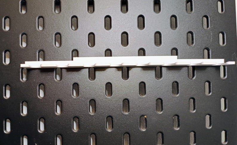 May include: White plastic pegboard hook strip with 10 hooks. The strip is mounted on a black pegboard.