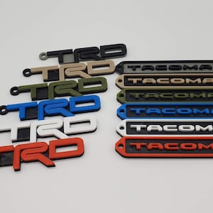 May include: A set of 10 keychains featuring the words "TRD" and "TACOMA" in various colors, including black, white, red, blue, green, and tan. The keychains are made of plastic and have a 3D printed look.