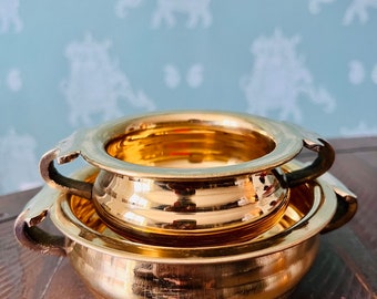 May include: Three nested, shiny gold-colored bowls with handles. The bowls are stacked on top of each other, with the smallest bowl at the top. The bowls are sitting on a wooden surface.