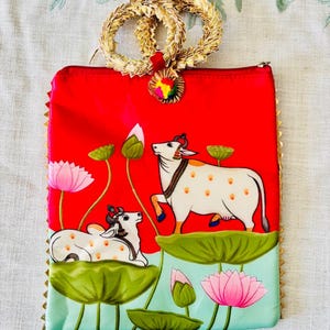 Pode incluir: Uma clutch vermelha com uma al&ccedil;a e acabamento dourados, apresentando uma cena pintada de vacas brancas, flores de l&oacute;tus e folhas de nen&uacute;far verdes. A clutch tem um fecho de correr e um pequeno charme colorido.