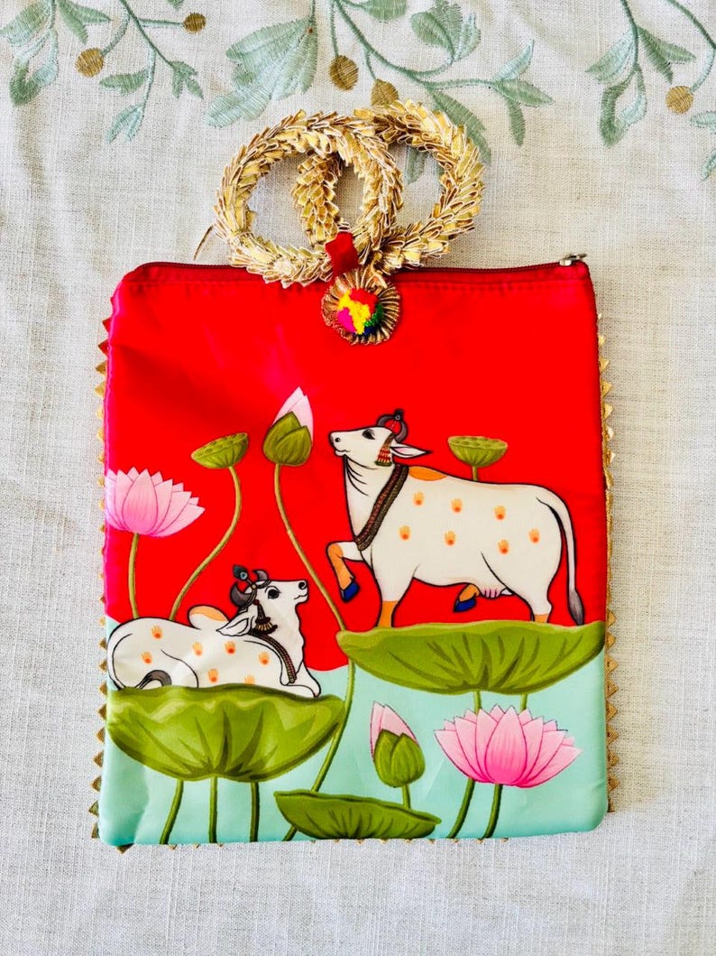 Pode incluir: Uma clutch vermelha com uma al&ccedil;a e acabamento dourados, apresentando uma cena pintada de vacas brancas, flores de l&oacute;tus e folhas de nen&uacute;far verdes. A clutch tem um fecho de correr e um pequeno charme colorido.