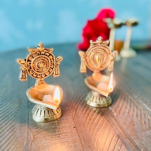 May include: Two gold metal oil lamps with intricate designs and lit wicks. The lamps are on a wooden surface.