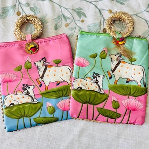 Pode incluir: Duas bolsas de m&atilde;o coloridas, uma rosa e outra azul-turquesa. Ambas apresentam uma ilustra&ccedil;&atilde;o de vaca com flores e folhas de l&oacute;tus. Cada bolsa tem uma al&ccedil;a tran&ccedil;ada e acabamentos dourados.