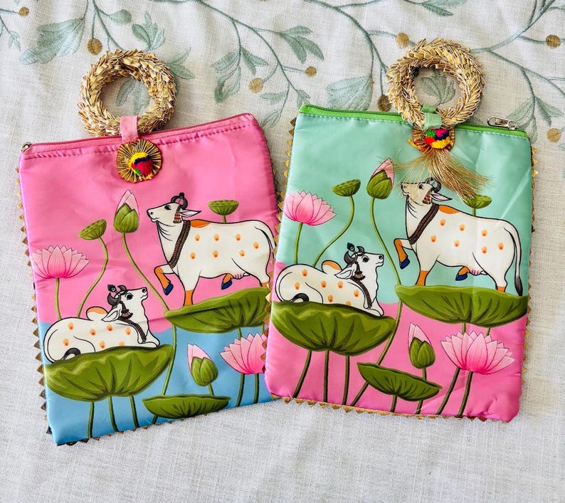 Pode incluir: Duas bolsas de m&atilde;o coloridas, uma rosa e outra azul-turquesa. Ambas apresentam uma ilustra&ccedil;&atilde;o de vaca com flores e folhas de l&oacute;tus. Cada bolsa tem uma al&ccedil;a tran&ccedil;ada e acabamentos dourados.