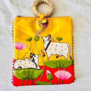 Pode incluir: Bolsa de m&atilde;o amarela com um design pintado &agrave; m&atilde;o com duas vacas brancas com manchas marrons, flores de l&oacute;tus e folhas de nen&uacute;far verdes. A bolsa tem uma al&ccedil;a tecida e uma borla com detalhes coloridos. As bordas s&atilde;o adornadas com uma borda decorativa.