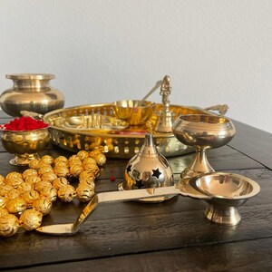 May include: A traditional Indian brass puja thali set with a variety of items, including a small bowl with red powder, a round tray, a small incense burner, a diya, and a set of golden beads.