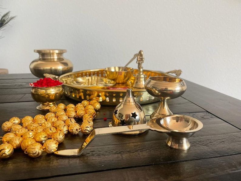 May include: A traditional Indian brass puja thali set with a variety of items, including a small bowl with red powder, a round tray, a small incense burner, a diya, and a set of golden beads.