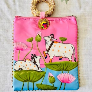Pode incluir: Bolsa clutch rosa com al&ccedil;a de palha tecida &agrave; m&atilde;o e bot&atilde;o decorativo. A bolsa apresenta uma pintura de duas vacas brancas com manchas laranja, flores de l&oacute;tus e nen&uacute;fares. As bordas s&atilde;o douradas.