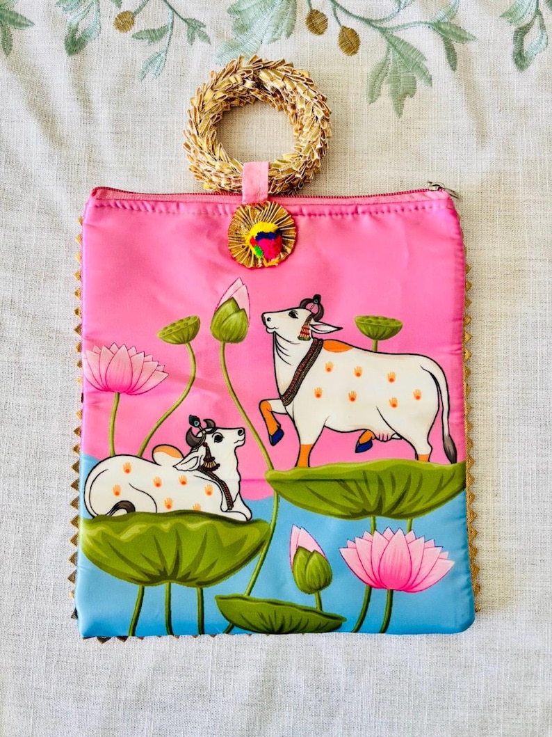 Pode incluir: Bolsa clutch rosa com al&ccedil;a de palha tecida &agrave; m&atilde;o e bot&atilde;o decorativo. A bolsa apresenta uma pintura de duas vacas brancas com manchas laranja, flores de l&oacute;tus e nen&uacute;fares. As bordas s&atilde;o douradas.