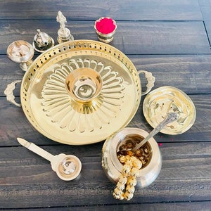 May include: A brass puja thali set with a large round plate, two small bowls, a spoon, and a small pot with a lid. The set is arranged on a wooden surface.