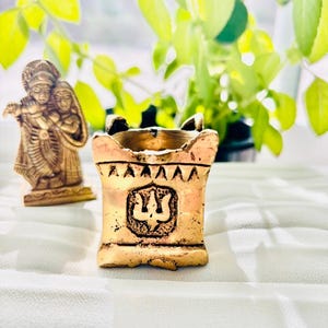May include: A small, gold-colored metal temple with a trident symbol on the front. The temple is sitting on a white surface.