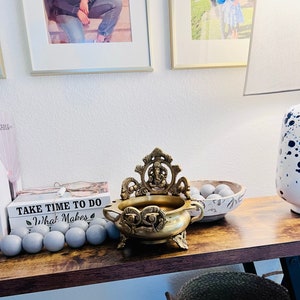 May include: A gold metal incense burner with an ornate design featuring a deity figure. The burner is sitting on a wooden table with a bowl of gray decorative balls and a stack of books with the text "Take Time To Do What Makes You Happy".