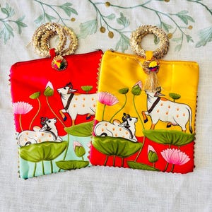 Pode incluir: Duas bolsas de m&atilde;o coloridas, uma vermelha e outra amarela. Cada uma apresenta um design pintado &agrave; m&atilde;o de vacas brancas com manchas marrons, flores de l&oacute;tus e folhas de nen&uacute;far. As bolsas t&ecirc;m al&ccedil;as e borlas douradas.