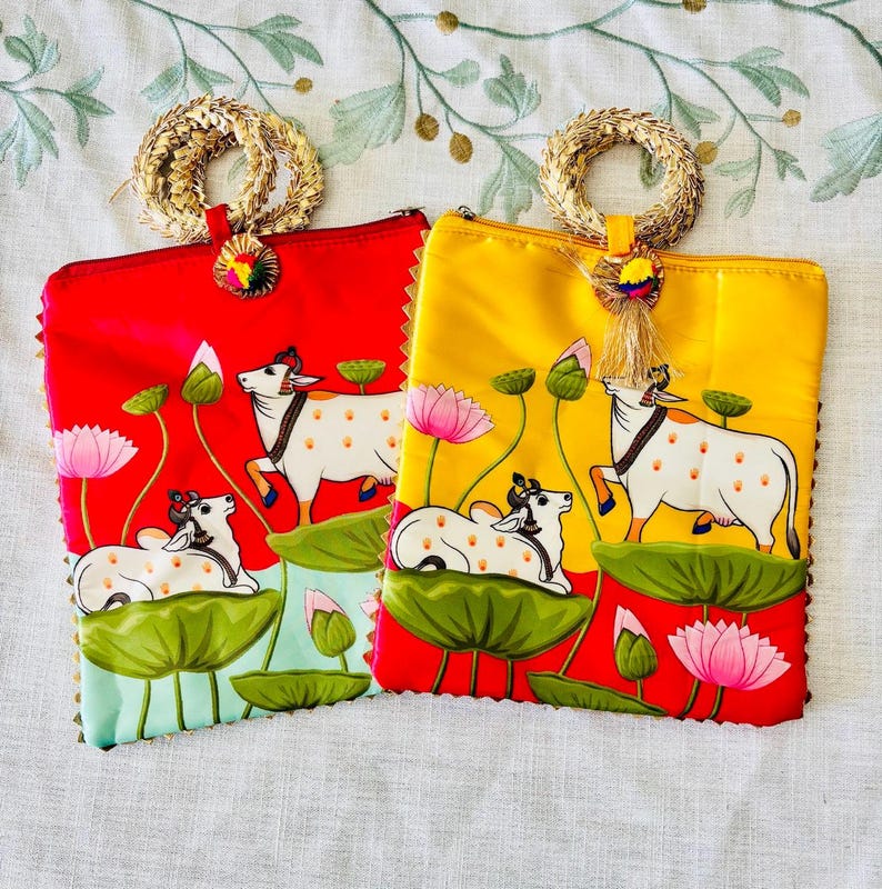 Pode incluir: Duas bolsas de m&atilde;o coloridas, uma vermelha e outra amarela. Cada uma apresenta um design pintado &agrave; m&atilde;o de vacas brancas com manchas marrons, flores de l&oacute;tus e folhas de nen&uacute;far. As bolsas t&ecirc;m al&ccedil;as e borlas douradas.