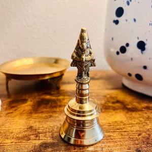 May include: A small, gold-colored bell with a detailed, ornate design. The bell has a rounded base and a slender, pointed top with a small, detailed figure.