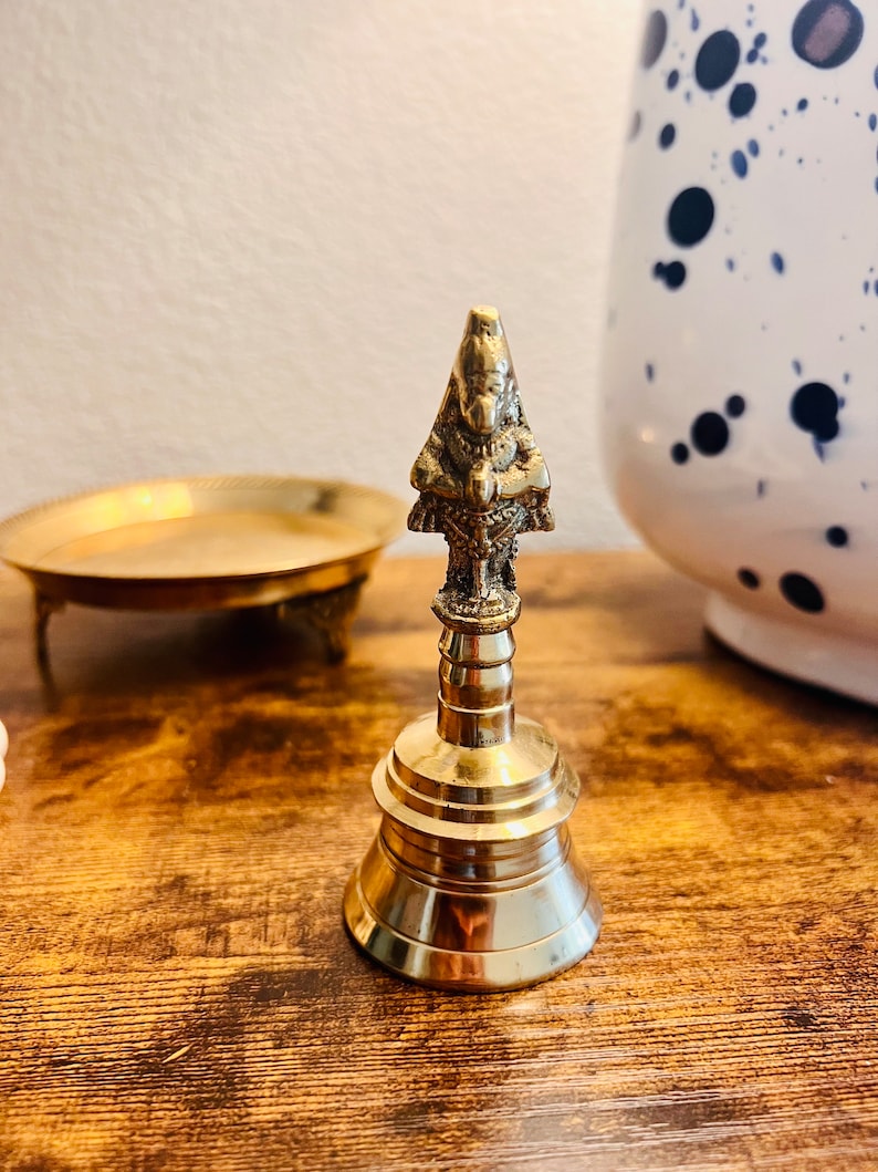 May include: A small, gold-colored bell with a detailed, ornate design. The bell has a rounded base and a slender, pointed top with a small, detailed figure.