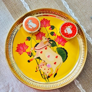 May include: A decorative yellow tray with a gold rim, featuring a painted design of a cow and calf with pink hearts and lotus flowers. Two small round containers with cow illustrations are also on the tray.
