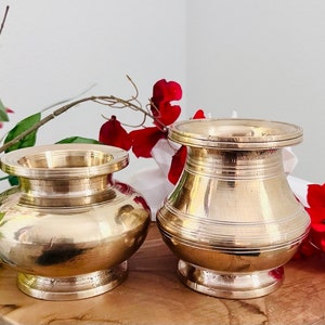 May include: Two gold metal bowls, one larger than the other, with a slightly flared lip. The bowls are sitting on a wooden surface with red flowers in the background.