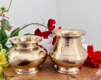May include: Two gold metal bowls, one larger than the other, with a slightly flared lip. The bowls are sitting on a wooden surface with red flowers in the background.