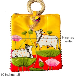 Pode incluir: Uma bolsa amarela, com 25 cm de altura e 23 cm de largura, com uma al&ccedil;a tecida. A bolsa apresenta uma pintura de duas vacas brancas com manchas marrons, flores de l&oacute;tus e folhas de nen&uacute;far verdes. A bolsa tem uma borda vermelha.