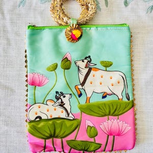 Pode incluir: Uma bolsa retangular com al&ccedil;a tecida e ilustra&ccedil;&atilde;o colorida. A bolsa apresenta um fundo azul claro e rosa com uma pintura de duas vacas brancas, flores de l&oacute;tus e folhas de nen&uacute;far verdes. A bolsa &eacute; adornada com dourado e tem um z&iacute;per verde.